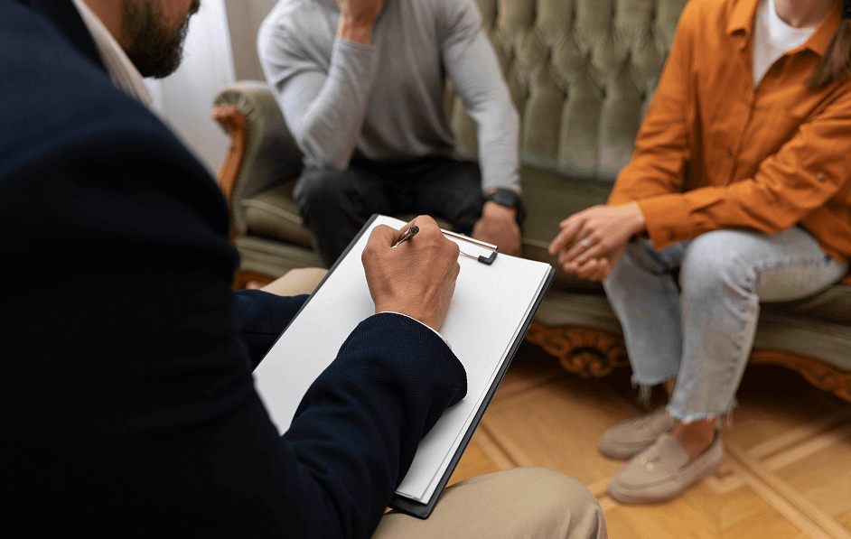 Person holding a clipboard and pen, listening to two individuals seated on a couch during a counseling session.